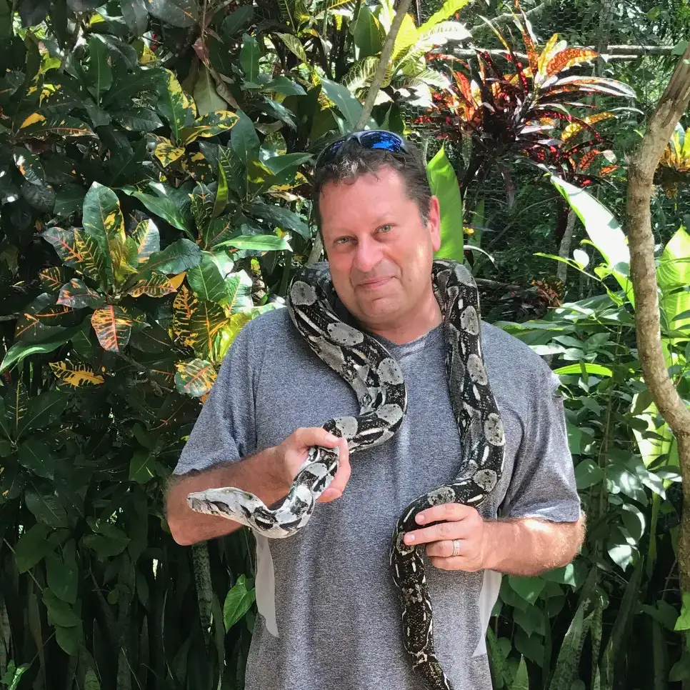 a man in a jungle setting wih a large snake handing around his shoulders.