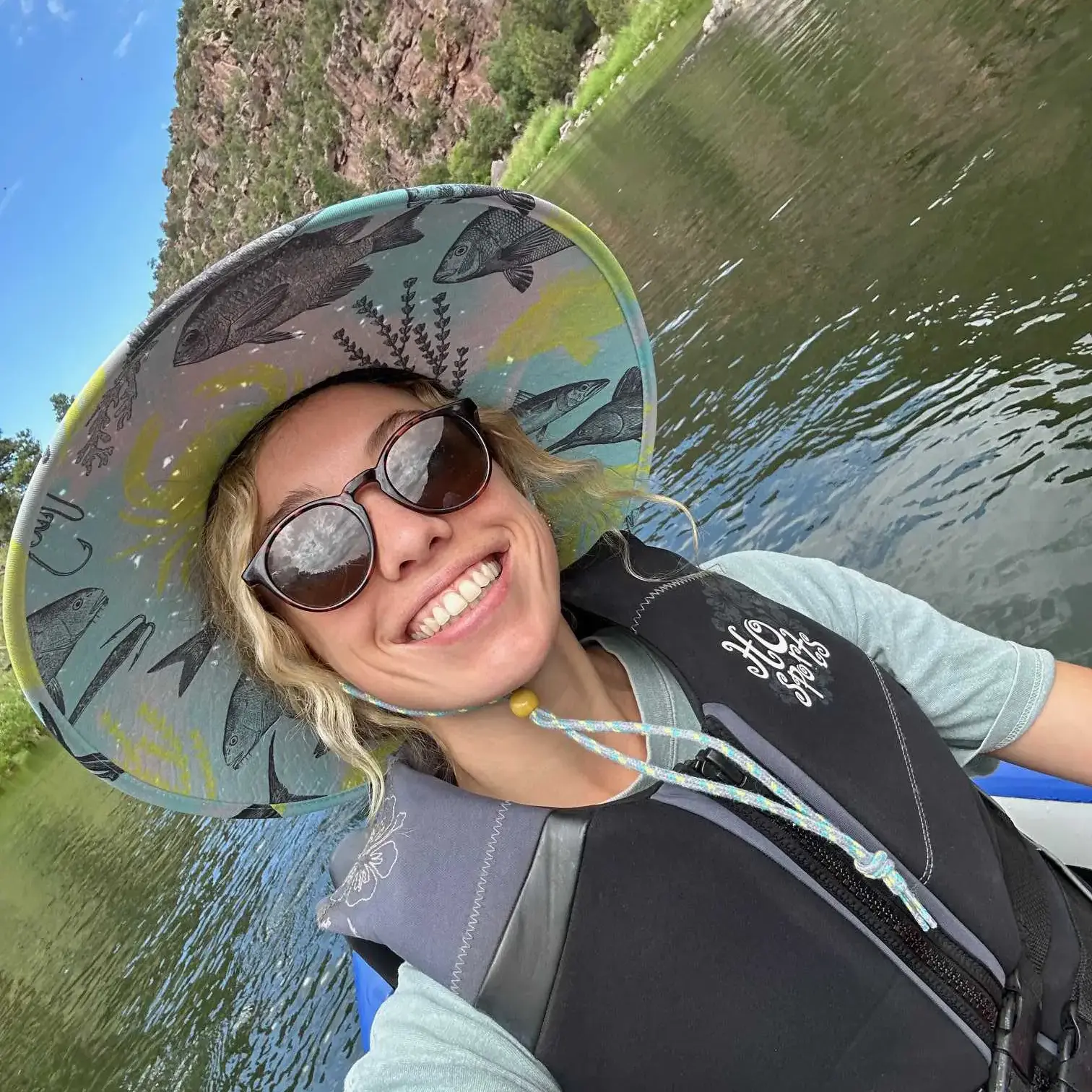 a blonde woman wearing a life jacket, sunglasses, and a colorful sun hat.